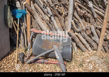 Ansammlung von Gegenständen an einem rustikalen Zaun mit Ästen und Holzstämmen, grauem Mopp, blauem abgenutzten alten Besen und Metallteilen auf dem Schmutzboden, Su Stockfoto