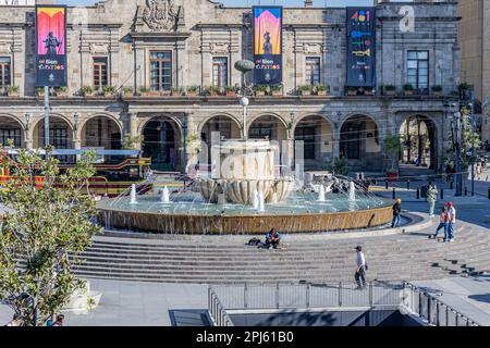 Guadalajara, Jalisco Mexiko. 4. Februar 2023. Brunnen an der Plaza de los Laureles mit Personen, die sitzen und spazieren gehen, Busse, Bäume und Stadtpala besichtigen Stockfoto