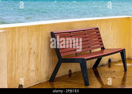 Leere hölzerne Bank, nass von Meerwasser neben einem Zaun an der Strandpromenade, Meer im Hintergrund, blaues Wasser ohne Wellen, ruhiger und bewölkter Tag in La Paz, Stockfoto