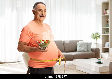 Ein erwachsener Mann in Sportbekleidung hält zu Hause einen Salat und misst die Taille im Wohnzimmer Stockfoto