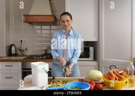 Glückliche Erwachsene Frau, die Gurken zum Mittagessen abzieht und organische Lebensmittelabfälle in einem Kompost-Behälter kompostiert Stockfoto