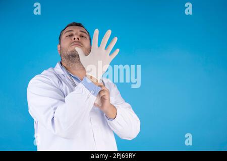 Ein Arzt mit einem ernsten Gesicht und einem Bart zieht chirurgische Handschuhe auf einem isolierten blauen Hintergrund an Stockfoto