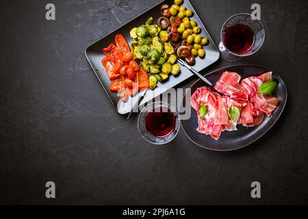 Prosciutto mit verschiedenen Vorspeisen mit einem Glas Rotwein auf schwarzem Hintergrund. Über gebratenes Gemüse, gesunde Snack Stockfoto