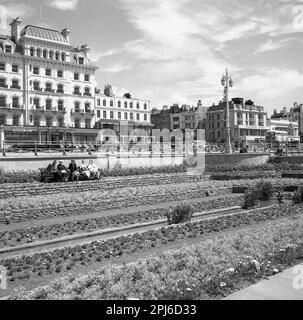 1950er, historischer Blick aus dieser Ära von Menschen, die auf Bänken in den Gärten am Meer sitzen, in Hove, W. Sussex, Brighton, England, UK. In der Ferne befindet sich das Norfolk Hotel in der Straße dahinter. Es wurde 1865 in der Kings Rd erbaut und von Horatio Nelson Goulty entworfen, einem englischen Architekten und einer wichtigen Figur in Brighton zu dieser Zeit. Auf dem Bild sind auch das Salisbury Hotel und die Tudor Lounge Bar zu sehen. Stockfoto