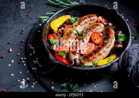 Gegrillte Würstchen und Gemüse mit Kräutern in einer eisernen Pfanne auf schwarzem Hintergrund Stockfoto