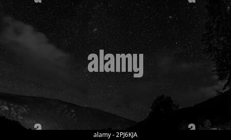 Schwarzweißfoto eines Sternenhimmels bei Nacht über einem Berg und einem Wald mit Wolken in den Bäumen des Altai in Sibirien. Stockfoto