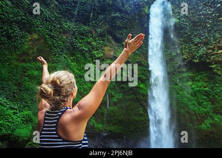 Junge, glückliche Frau im Touristenlager, die Hände ausstreckt und die Natur und den Süßwasserfall genießt, die durch den Bali-Regenwald reisen. Tagestour durch die Natur, Wandern Stockfoto