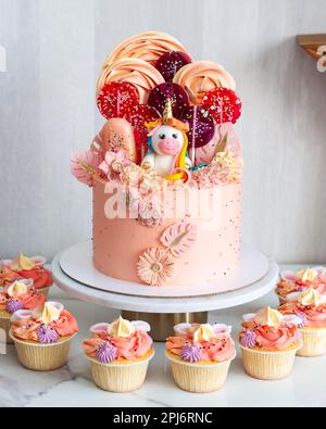 Süßer rosa Geburtstagskuchen für ein kleines Mädchen mit einem Einhorn und Baiser-Wirbeln. Lutscher, Schokoladenblumen und Cupcakes auf grauem Hintergrund Stockfoto