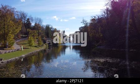 Wunderschön gestalteter Park mit dekorativem Brunnen in der Mitte des Sees, Bäume und große Steine an Ufern an sonnigen Herbsttagen. Fliegen über Wasser mit heruntergefallenen Blättern und blauem Himmel. Viele Touristen Stockfoto