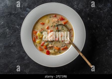 Frische Fischsuppe mit Karotten, Kartoffeln, Tomaten und roten Paprika auf einem weißen Teller auf schwarzem Hintergrund, Nahaufnahme. Das leckere Abendessen besteht aus einer Sahne Stockfoto