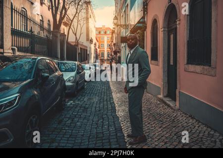 Ein Mann in einem stylischen grau-grünen Anzug und einer Sonnenbrille steht auf einem schmalen Bürgersteig, Hände in den Taschen. Gebäude umzingeln ihn, geparkte Autos sind an der Reihe Stockfoto