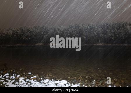 Nachts fällt weißer Schnee in der Dunkelheit in einem Lichtstrahl vor dem Hintergrund des Flusses Katun und im Herbst in Altai Wälder. Lange Belichtung. Stockfoto