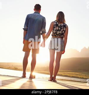 Liebe und Sonnenuntergänge. Rückblick auf ein liebevolles junges Paar, das bei Sonnenuntergang auf einem Dock steht. Stockfoto