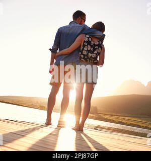 Liebe kann nicht besser werden als das. Rückblick auf ein liebevolles junges Paar, das bei Sonnenuntergang auf einem Dock steht. Stockfoto