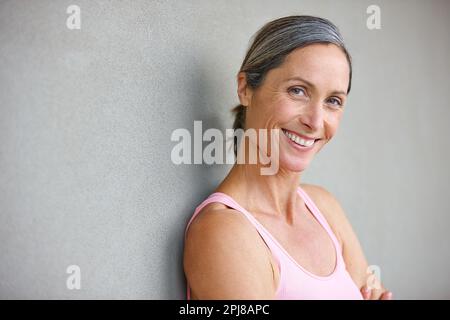 Sie ist in guter Verfassung. Porträt einer attraktiven, reifen Frau in Turnkleidung, die sich gegen eine graue Wand lehnt. Stockfoto
