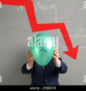 Ein Geschäftsmann hielt einen Luftballon mit einem traurigen Gesicht und einer Abbildung eines herunterfallenden Diagramms auf hellgrauem Hintergrund. Rezessionskonzept der Wirtschaft Stockfoto