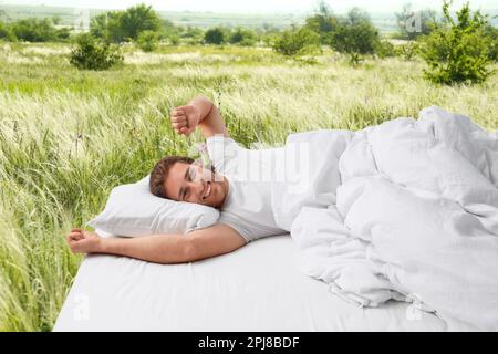 Glücklicher Mann auf dem Bett mit weichen Kissen und wunderschöner Aussicht auf die grüne Wiese im Hintergrund. Gut schlafen - gesund bleiben Stockfoto