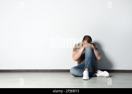 Trauriger junger Mann, der auf dem Boden neben der weißen Wand im Haus sitzt, Platz für Text Stockfoto