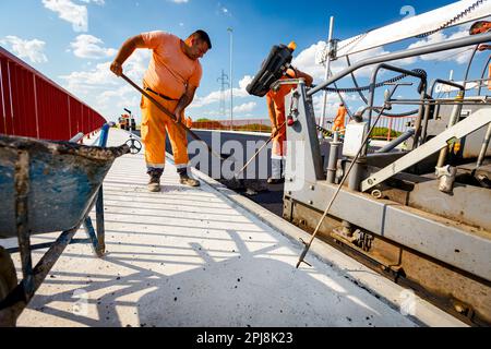 Ansicht auf der Maschine zum Verlegen von Asphalt, Verteilen einer heißen Asphaltschicht auf vorbereitetem Boden, an dem einige Arbeiter arbeiten. Stockfoto