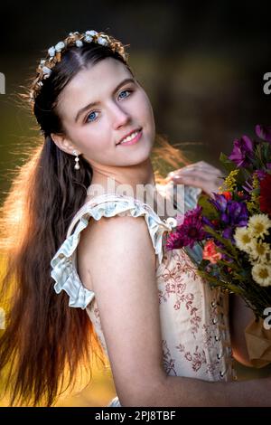 Wunderschöne junge Frau in einem Cottage-Kleid und einem knöchernen Oberteil, die einen Blumenstrauß in der Hand hält | Hintergrundbeleuchtung | Wald Stockfoto