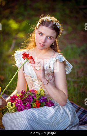 Junge Frau mit knöpftem Oberteil und Kleid, sitzt auf einer Decke und hält einen Blumenstrauß in der Hand | Haarband mit Blume | warme Farben Stockfoto