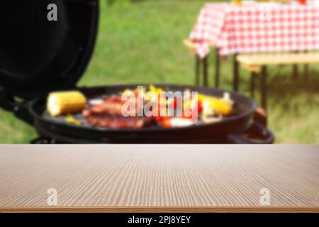 Leerer Holztisch und verschwommener Blick auf leckeres Essen auf dem modernen Barbecue-Grill im Freien Stockfoto