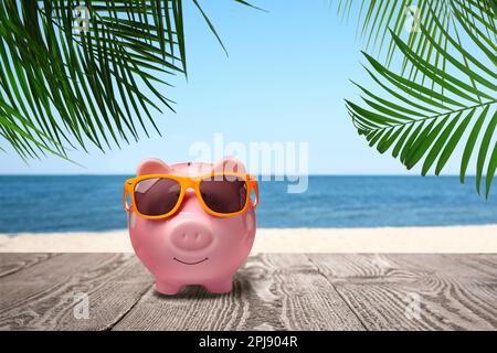 Sparen Geld für den Sommerurlaub. Sparschwein mit Sonnenbrille auf Holzfläche in der Nähe von Sandstrand und Meer Stockfoto