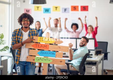 Ein junger männlicher Büroangestellter hält ein Schild mit Firmenlogans und posiert für ein Foto in einer angenehmen Atmosphäre im Büro mit seinen Kollegen. E Stockfoto