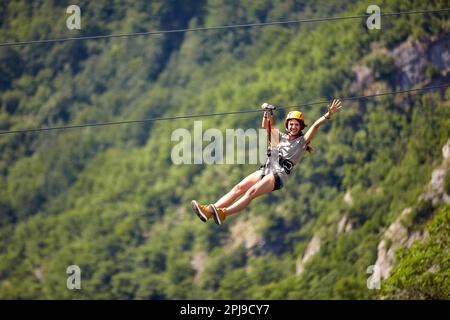 Glückliche junge, fröhliche weibliche Tourist in lässiger Kleidung, die auf einer Seilrutsche im Wald reitet. Zipline-Tour mit selektivem Fokus vor verschwommenem Hintergrund. Holi Stockfoto