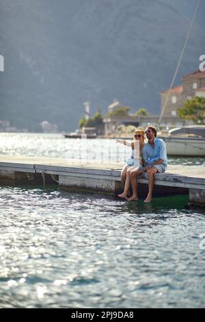 Ein junges Paar sitzt mit den Beinen im Wasser auf dem Dock am Meer und genießt die Aussicht an einem wunderschönen sonnigen Tag. Liebe, Beziehung, Stockfoto