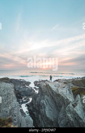 Furchtloser Mann mit Rucksack, der den Sonnenuntergang über dem Atlantik in Porto Covo, Portugal, beobachtet. Ein Wanderer steht am Rand einer Klippe und blickt in h Stockfoto