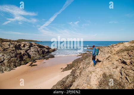 Backpacker wandern entlang eines Felsens und über einen Sandstrand auf dem markierten Fisherman Trail an der Atlantikküste in der Nähe von Porto Covo, Portugal. Rota Vicentina. Stockfoto