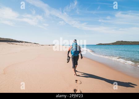 Ein schwarzhaariger gutmütiger Mann mit Rucksack und Schuhen in den Händen geht am Strand von Praia da Ilha do Pessegueiro entlang, am Atlantik in der Nähe Stockfoto