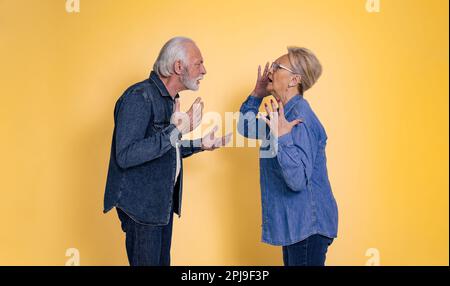 Seitlicher Blick auf ein wütendes älteres Paar in blauen Jeans, das sich streitet und anschreit, während es von Angesicht zu Angesicht isoliert über gelbe Hintergräben steht Stockfoto