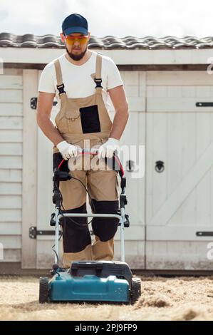 Der Mensch-Gärtner, der Rasen mit einem Vertikutierer erscharft, erscharft den Rasen und entfernt altes Gras Stockfoto