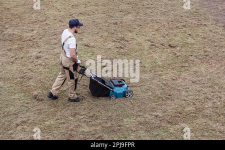 Der Mensch-Gärtner, der Rasen mit einem Vertikutierer erscharft, erscharft den Rasen und entfernt altes Gras Stockfoto