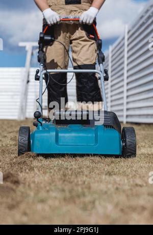 Der Mensch-Gärtner, der Rasen mit einem Vertikutierer erscharft, erscharft den Rasen und entfernt altes Gras Stockfoto