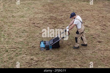 Der Mensch-Gärtner, der Rasen mit einem Vertikutierer erscharft, erscharft den Rasen und entfernt altes Gras Stockfoto