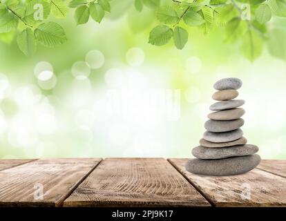 Gestapelte Steine auf einem Holztisch unter grünen Blättern vor verschwommenem Hintergrund, Platz für Text. Zen-Konzept Stockfoto