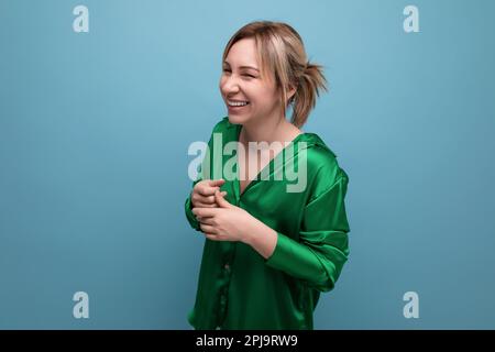 Attraktive blonde junge Frau in grünem Seidenhemd, die auf blauem Hintergrund lacht Stockfoto