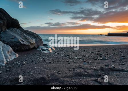 Playa de Ajabo Strand mit schwarzem vulkanischem Sand und Felsen während des Sonnenuntergangs. Foto mit langer Belichtung. Fischersilhouette am Pier. Stockfoto