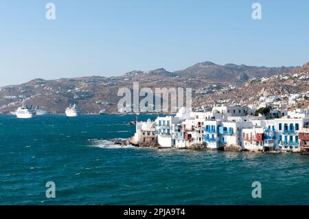 Panoramablick auf das Dorf Chora auf der griechischen Insel Mykonos, im Hintergrund die felsigen Hügel und verankerten Kreuzfahrtschiffe. Mediterranes Vakuum Stockfoto