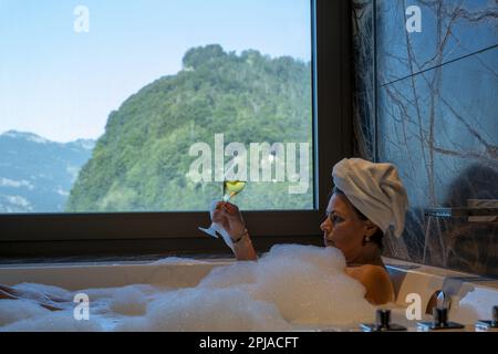 Frau liegt in einer Badewanne mit einem Glas Champagner und genießt den Fensterblick über den Berg an einem sonnigen Sommertag in Burgenstock, Nidwalden, Switzerlan Stockfoto