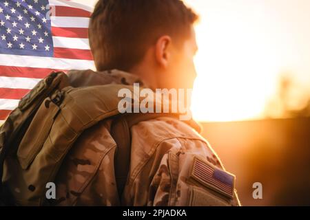 Großartig für den 4. Juli oder Memorial Day. Rückansicht eines US-Soldaten mit Blick auf den Sonnenuntergang und einer US-Flagge. Stockfoto