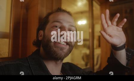 Bärtiger junger Mann in POV mit Telefon und Lachen während eines Videogesprächs auf einem gelb beleuchteten Holzbalkon Stockfoto