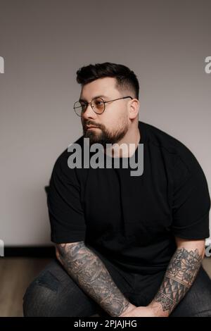 Ein ruhiger, bärtiger und tätowierter Mann, der im Studio auf einem Stuhl sitzt Stockfoto