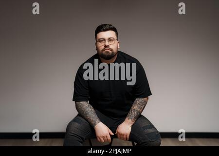 Ein ruhiger, bärtiger und tätowierter Mann, der im Studio auf einem Stuhl sitzt Stockfoto