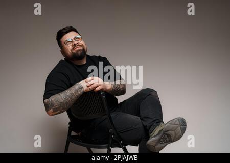 Ein ruhiger, bärtiger und tätowierter Mann, der im Studio auf einem Stuhl sitzt Stockfoto