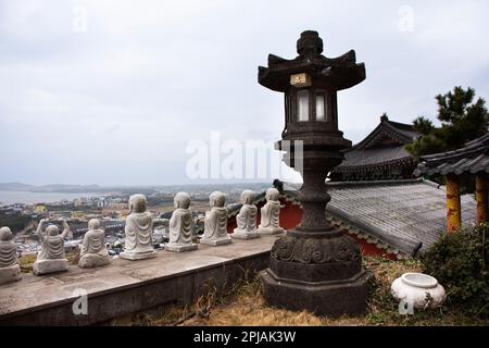 Steinlaterne im antiken koreanischen Stil und kleine buddha-Statue am Aussichtspunkt im Sanbangsa-Tempel mit Blick auf die landschaftlich reizvolle Insel Jeju und das Stadtbild Seogwipo CI Stockfoto