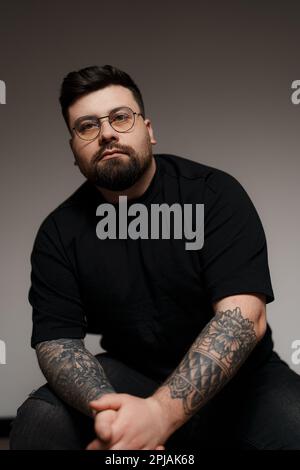 Ein ruhiger, bärtiger und tätowierter Mann, der im Studio auf einem Stuhl sitzt Stockfoto